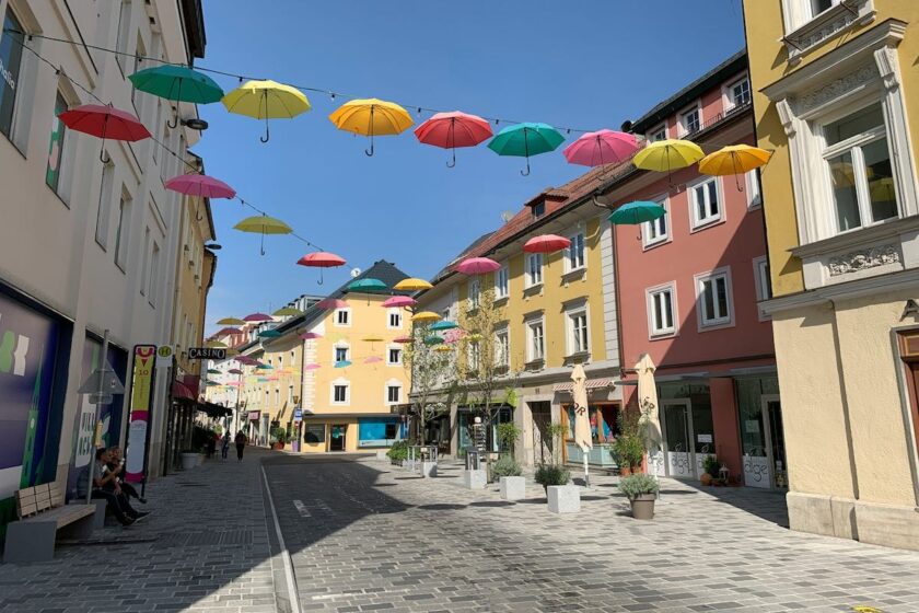 Decorazione urbana nel centro storico di Villach, con ombrelli colorati appesi tra i palazzi in una via pedonale. Un’installazione vivace e creativa che valorizza l’estate in città.