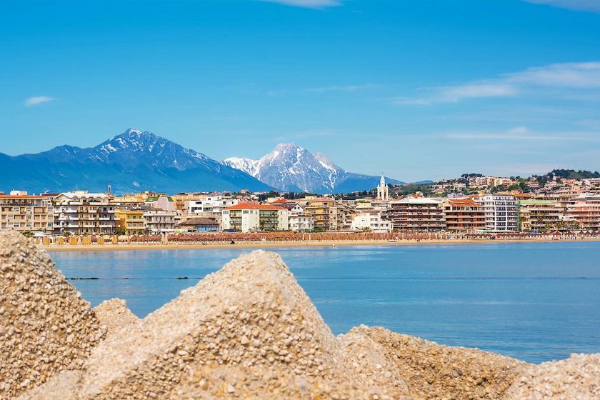 Vista del lungomare di Pescara con gli ombrelloni sulla spiaggia, il campanile della chiesa del Divino Amore e il Gran Sasso sullo sfondo.